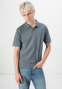 Camisa polo gris con un cuello corto y una tapeta de tres botones; combinada con jeans azul claro. Tejido suave con un detalle de logo mínimo.