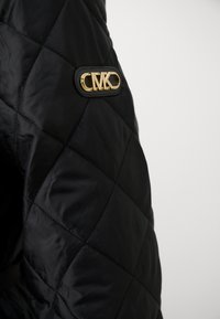Black quilted jacket fabric with diamond pattern, featuring a gold logo patch on the upper sleeve. Smooth texture and tailored design.