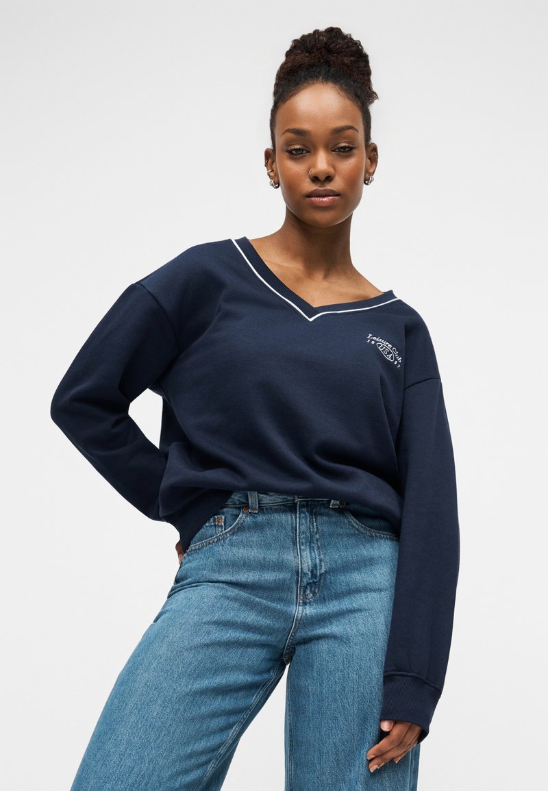 Marineblauwe sweatshirt met een V-hals en witte bies, met een logo op de borst. Gecombineerd met lichtblauwe spijkerbroek. Zachte stof en een ontspannen pasvorm.