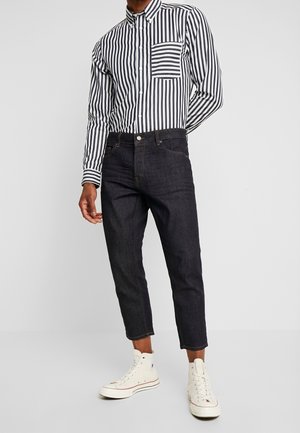 Man draagt zwarte afgekorte jeans, een zwart-wit verticaal gestreept overhemd met knopen en witte hoge sneakers.