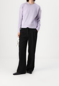 Person, der einen helllila Pullover, weite schwarze Hosen, schwarze Schuhe trägt und eine kleine weiße Handtasche hält, steht vor einem weißen Hintergrund.