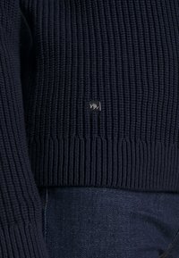 Pull en maille côtelée bleu marine avec un petit écusson logo carré en bas devant, associé à un jean en denim bleu foncé.