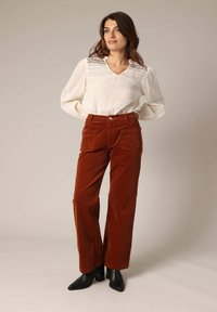 Blouse crème avec motif texturé et décolleté en V, associée à un pantalon large en velours côtelé couleur rouille et des bottes à talons noires.