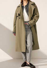 Olijfgroene trenchcoat met een losse pasvorm, dubbele knopen en een brede kraag, laag over een grijze denimjack en lichtgrijze broek.