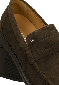 Mocassin en daim marron avec détails cousus, petit logo de la marque argenté sur la tige, et doublure intérieure en cuir beige.