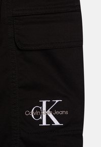 Černé látkové cargo kalhoty mají náprsní kapsu a vyšívané logo "Calvin Klein Jeans" v bílém provedení s klasickým písmem.