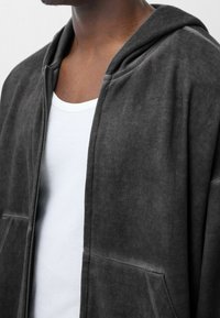 Primer plano de una persona que lleva una sudadera con capucha de cremallera gris oscuro sobre una camiseta blanca de cuello redondo, mostrando el cuello y la parte superior del pecho.