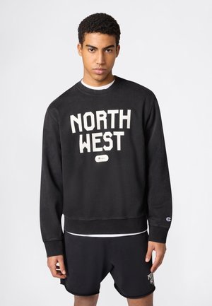 Młody mężczyzna ubrany w czarną bluzę i szorty z napisem "NORTH WEST", stojący na tle jednolitego jasnoszarego tła.