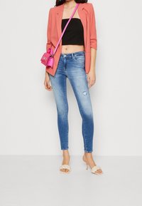Jeans ajustados azules con un acabado desgastado, una blusa de tiro corto negra, una blazer coral y un pequeño bolso cruzado rosa. Unas sandalias de tacón beige completan el look.