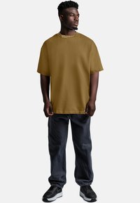 Maglietta oversize marrone con collo a giro, abbinata a pantaloni grigio scuro. Presenta una texture liscia e un design semplice con accenti minimi.
