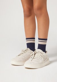 Baskets blanches texturées avec une semelle en caoutchouc, accompagnées de chaussettes bleu marine avec deux rayures blanches près du haut. Les pieds sont positionnés ensemble.