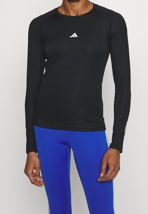 Persoon draagt een aansluitend zwart shirt met lange mouwen en een klein wit logo op de borst en felblauwe sportlegging.