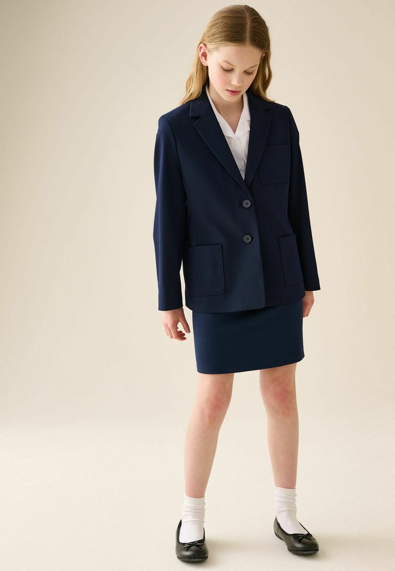 Jeune femme vêtue d’un blazer bleu marine et d’une jupe assortie, chemise blanche, chaussettes blanches et ballerines noires, debout devant un fond beige uni.