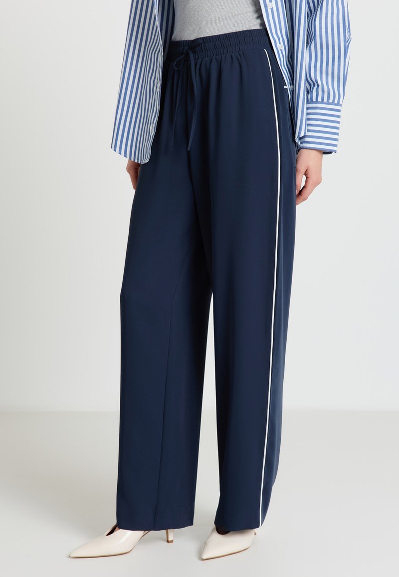 Donna che indossa pantaloni larghi blu navy con striscia laterale bianca, tacchi bianchi a punta, top grigio e camicia a righe blu e bianche.