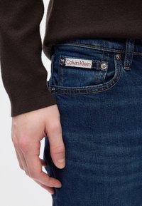 Tumši zili džinsi ar vieglu nodilumu un raksturīgo Calvin Klein etiķeti labajā kabatā. Roka balstās uz džinsu sāniem.