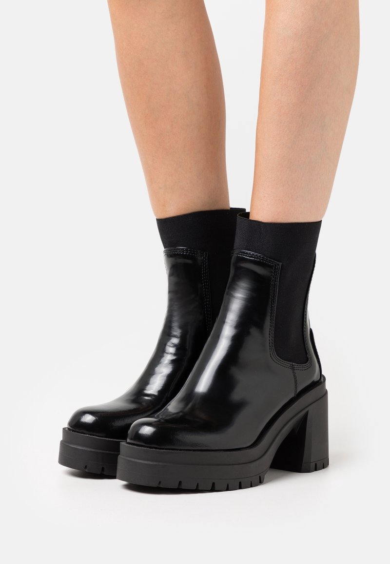 Bottines noires brillantes avec talons épais et panneaux en tissu extensible portées sur des jambes nues sur fond blanc.