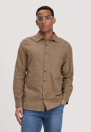 Junger Mann mit klarer Brille, einem braun karierten Button-up-Hemd und blauen Jeans, stehend mit einer Hand in der Tasche vor einfarbigem Hintergrund.