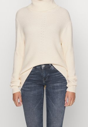 Pull en maille crème à col roulé associé à un jean slim bleu foncé, mains détendues le long du corps, fond clair uni.