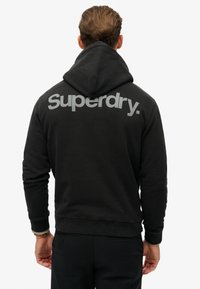Sudadera negra con un corte relajado, que presenta un gran logo gris de "Superdry" en la espalda, bolsillo tipo canguro y capucha ajustable.