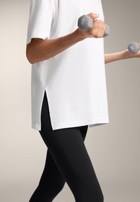 Hvid kortærmet skjorte med sidesplitter, parret med sorte leggings; individet holder grå 1 kg håndvægte i hver hånd.