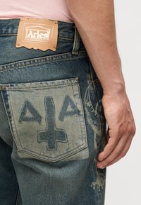 Primer plano de una persona que lleva vaqueros azules con un parche de cuero de la marca Aries y un símbolo oscuro impreso en el bolsillo trasero.