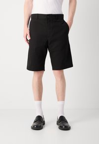 Pantaloni scurți negri din material, cu un design clasic, având închidere cu nasture și buzunare laterale, purtați cu șosete albe și pantofi loafer negri.