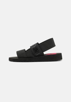 Sort flad sandal med to brede elastiske remme, én med hævet tekst med mærke, og en slingback rem på en ensfarvet baggrund.