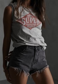 T-shirt sans manches gris avec un logo en forme de diamant rouge portant l'inscription "DRY SUPER GENUINE GOODS", accompagné d'un short en denim noir usé avec des bordures effilochées.