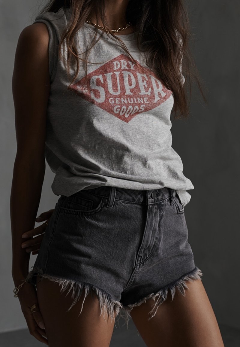 T-shirt sans manches gris avec un logo en forme de diamant rouge portant l'inscription "DRY SUPER GENUINE GOODS", accompagné d'un short en denim noir usé avec des bordures effilochées.