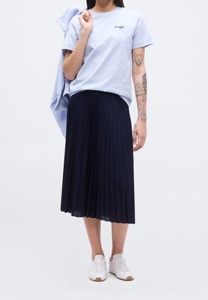 Vrouw die een lichtblauw T-shirt draagt, een zwarte geplooide midirok, witte sneakers, houdt een blauwe jas over haar schouder en toont tatoeages op haar rechterarm.