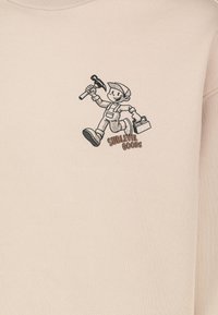 Beiger Pullover aus weichem Stoff mit einem aufgedruckten Charakter, der einen Hammer und einen Werkzeugkasten hält, mit dem Text "SUBLEVEL GOODS" darunter.