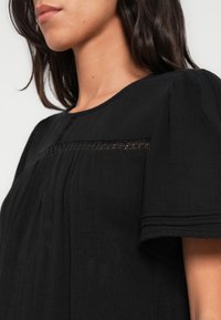 Μαύρο, ελαφρύ μπλουζάκι με κοντά φουσκωτά μανίκια, στρογγυλή λαιμόκοψη και οριζόντια δαντελένια λεπτομέρεια. Διαθέτει πιετέ υφή και απαλό ύφασμα.