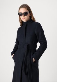 Marinblå ullrock med hög krage, bälte i midjan och sidofickor. Har en snygg design och len textur, lämplig för kallare väder.