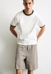 T-shirt blanche à manches courtes avec des finitions brunes, associée à un short beige. Le tissu semble lisse avec une coupe décontractée. Design simple.
