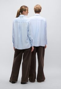 Ανοιχτό μπλε πουκάμισο με μακριά μανίκια και υφή, πτυχώσεις στην πλάτη και μανσέτες με κουμπιά, σε συνδυασμό με καφέ παντελόνι φαρδιάς γραμμής.