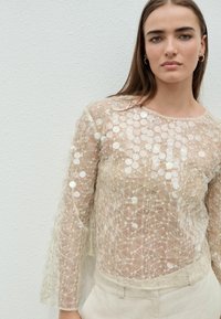Blouse transparente avec un design en réseau beige complexe, ornée d'accents circulaires blancs. Présente des manches longues évasées et un col rond.