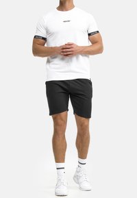 Mann steht mit gefalteten Händen und trägt ein weißes T-Shirt mit schwarzen Ärmelbündchen, schwarze Shorts, weiße Socken und weiße Sneaker.