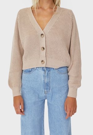 Femme portant un cardigan beige en tricot à boutons avec un décolleté en V et un jean bleu clair taille haute, visible des épaules aux hanches.
