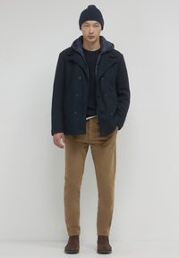 Cappotto doppiopetto navy con una finitura testurizzata, abbinato a un maglione scuro, pantaloni in velluto a coste color sabbia e stivaletti alla caviglia in suede marrone.