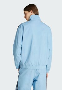 Lichte blauwe jas met een hoge kraag en elastische manchetten, met een relaxte fit en een gladde textuur. Zwarte zijaccenten zichtbaar.