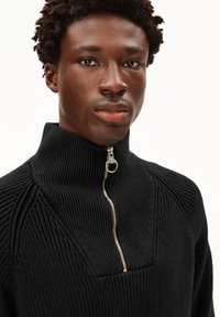 Junger schwarzer Mann, der einen schwarzen gerippten Strickpullover mit einem silbernen Halb-Reißverschluss-Kragen trägt und mit einem neutralen Gesichtsausdruck direkt in die Kamera schaut.
