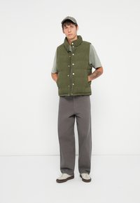 Vest căptușit de culoare verde măslin, cu guler înalt și nasturi tip snap, combinat cu un tricou gri deschis și pantaloni gri cu picioare largi, completat cu teniși albi.
