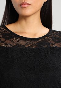 Mustat pitsi yläosa, jossa on läpinäkyvä kukkakuosi kaula-aukossa ja kiinteää kangasta alapuolella. Suunnittelussa on pyöreä kaula-aukko ja pehmeä tekstuuri.