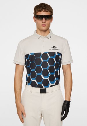 Polo shirt in off-white with a blue and black hexagon pattern. Features a classic collar, short sleeves, and logo detail. Worn with light-colored pants.
