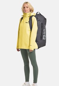 Gelbe wasserdichte Jacke, grauer Rucksack mit Logo, grüne Leggings und weiße Sneakers. Die Jacke hat einen hohen Kragen und eine verstellbare Kapuze.