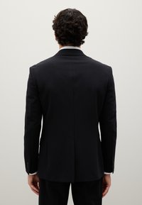 Giacca da abito nera slim-fit con rever a scollatura, apertura centrale e bottoni sulle maniche, vista da dietro su uno sfondo neutro.