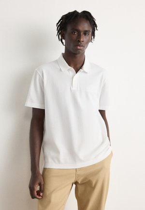 White polo shirt with a buttoned collar, crafted from soft material, featuring subtle logo embroidery, paired with beige pants.