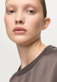 Close-up of a young person with light skin and brown hair, wearing a grey, ribbed crewneck top. Focus on soft features and neutral makeup.