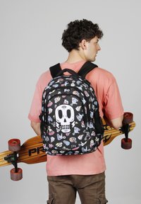 Mochila negra con patrones coloridos de calaveras y mariposas, que cuenta con correas ajustables y un bolsillo con cremallera en la parte frontal, sostenida por una persona con una patineta.