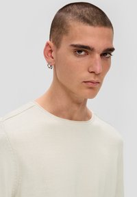 Helles beiges Langarmshirt mit Rundhalsausschnitt und genähten Details an den Schultern, getragen von einer Person mit kurzen Haaren und Ohrringen.